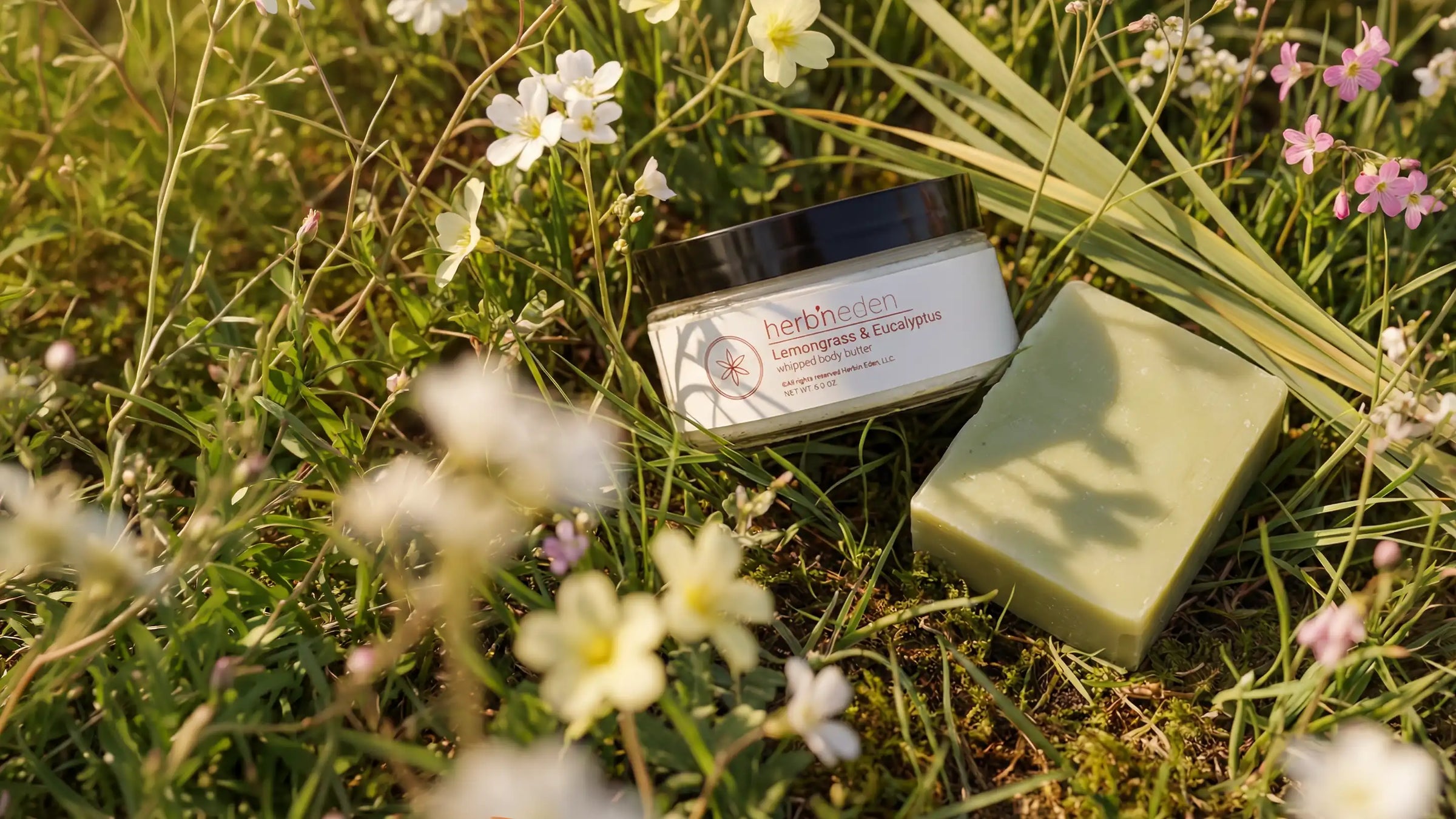 Herb’n Eden Lemongrass & Eucalyptus body butter and matching bar soap placed in grass among wildflowers, lit by warm natural sunlight.
