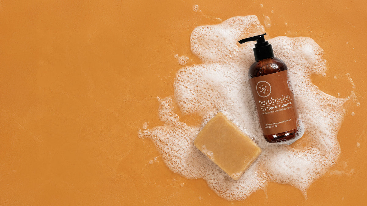 A bottle of Herb'neden Tea Tree & Turmeric wash with a black pump, placed on white foam next to a yellow soap bar on a solid orange background.